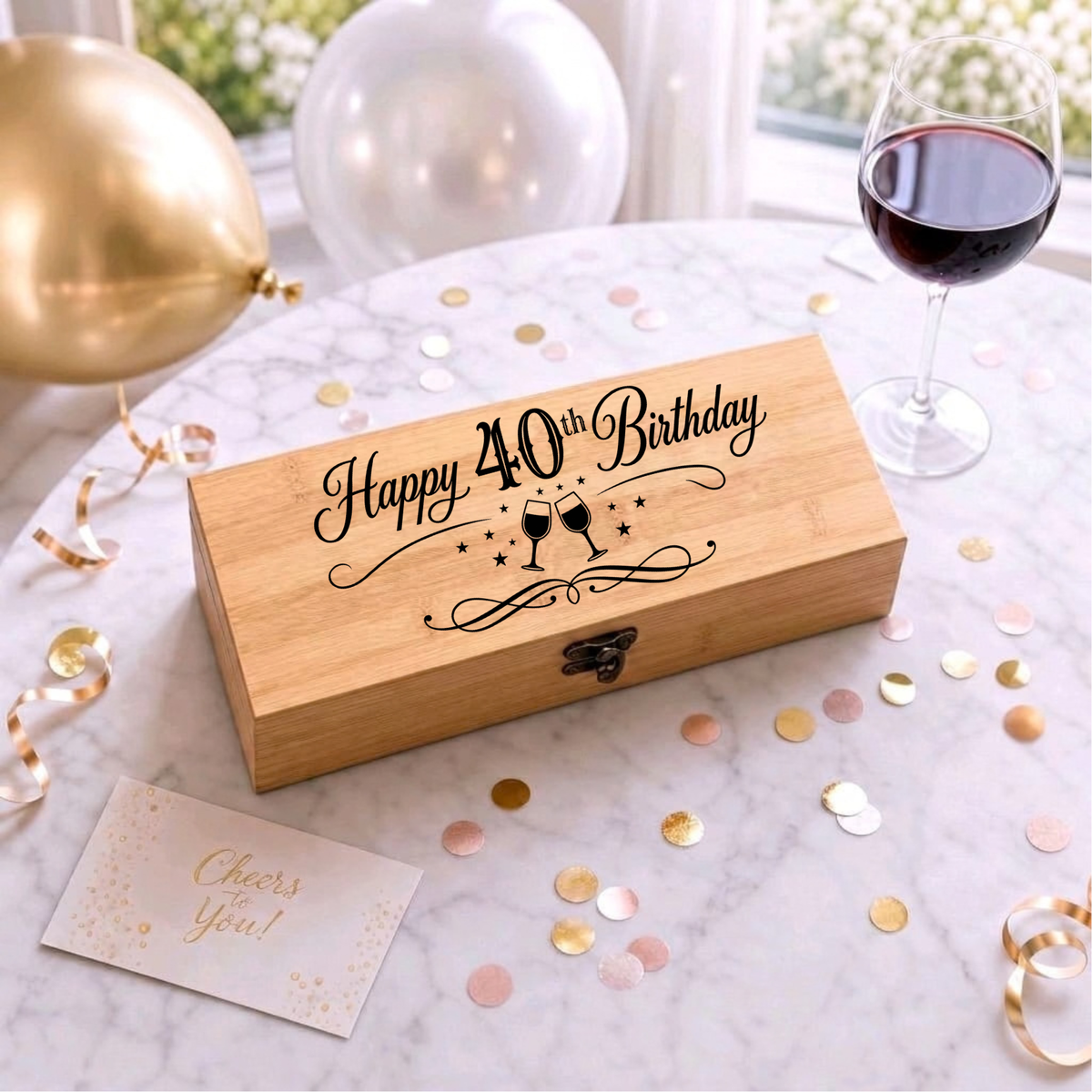 A wooden keepsake box engraved with 'Happy 40th Birthday' and a clinking glasses design, styled on a marble table with balloons, a glass of red wine, confetti, and a 'Cheers You!' card.