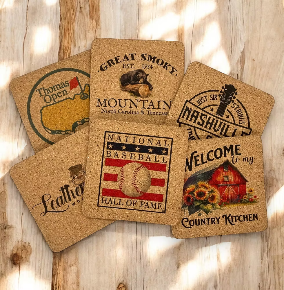 Five square cork coasters with rustic printed designs and text, arranged on a light wood surface with dappled sunlight and shadows.
