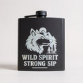 A matte black metal flask with a screw-top lid and carrying loop, featuring a white graphic of a wolf, mountains, trees, and the text 'WILD SPIRIT STRONG SIP' on the front, set against a plain light background.