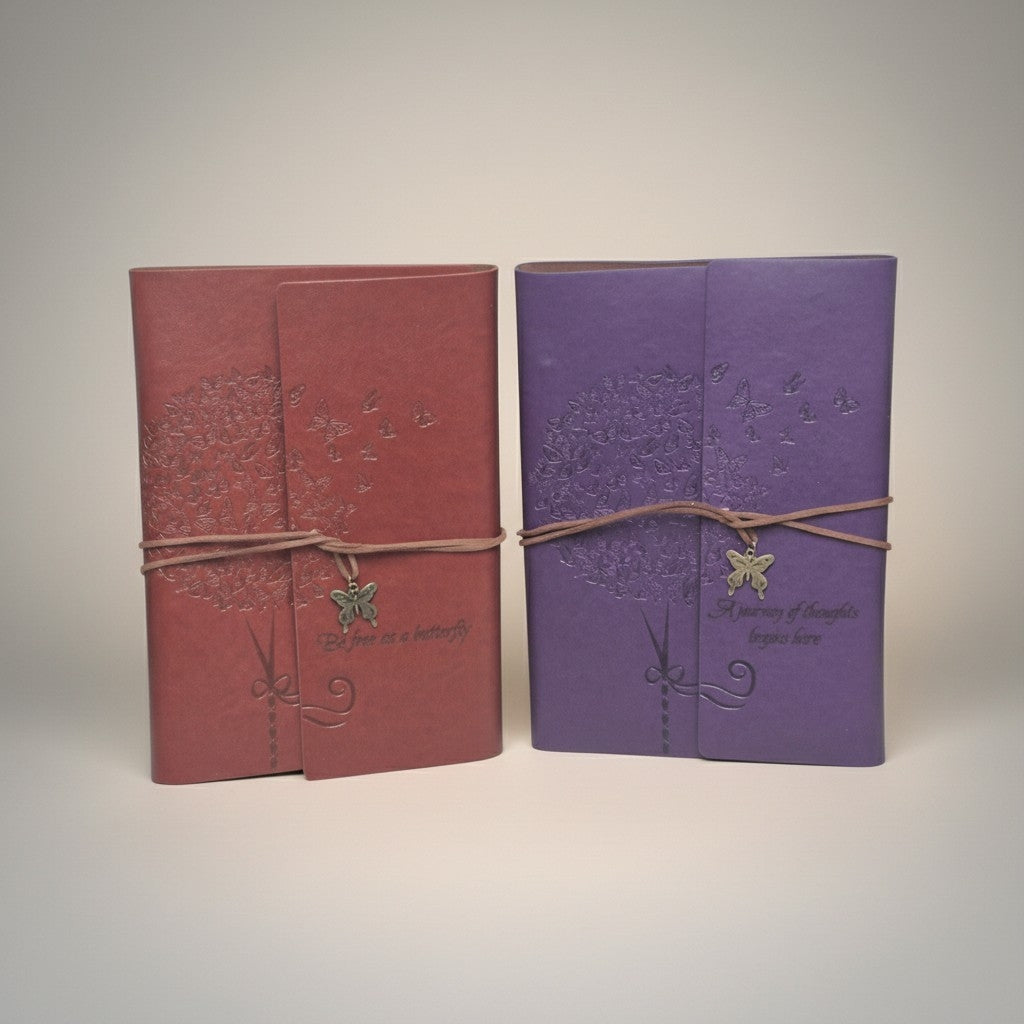 Two small hardcover journals or notebooks standing upright side by side on a light gray background. The left one is dusty rose pink with a brown twine wrap and a butterfly charm, and the right one is lavender purple with a matching twine wrap and butterfly charm.