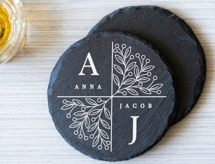 Two round black slate coasters on a light wood surface. The front coaster is personalized with a white cross dividing the design into four quadrants, featuring the names 'ANNA' and 'JACOB' with decorative botanical line art, alongside large letters 'A' and 'J'.
