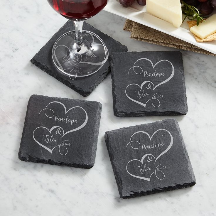 Four dark slate coasters with white engraved heart designs and the names 'Pendope & Tyler' are arranged on a white marble surface. A glass of red wine rests on one coaster, and cheese and grapes are visible in the background.