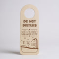 A novelty hanging tag or sign with a rounded top and circular hole for hanging, printed with the phrase 'DO NOT DISTURB RELAXING IN THE TUB' and a bathtub illustration. The tag appears to be a light natural wood or cardboard-like beige color with darker brown text and line art, shown against a plain light gray studio background.