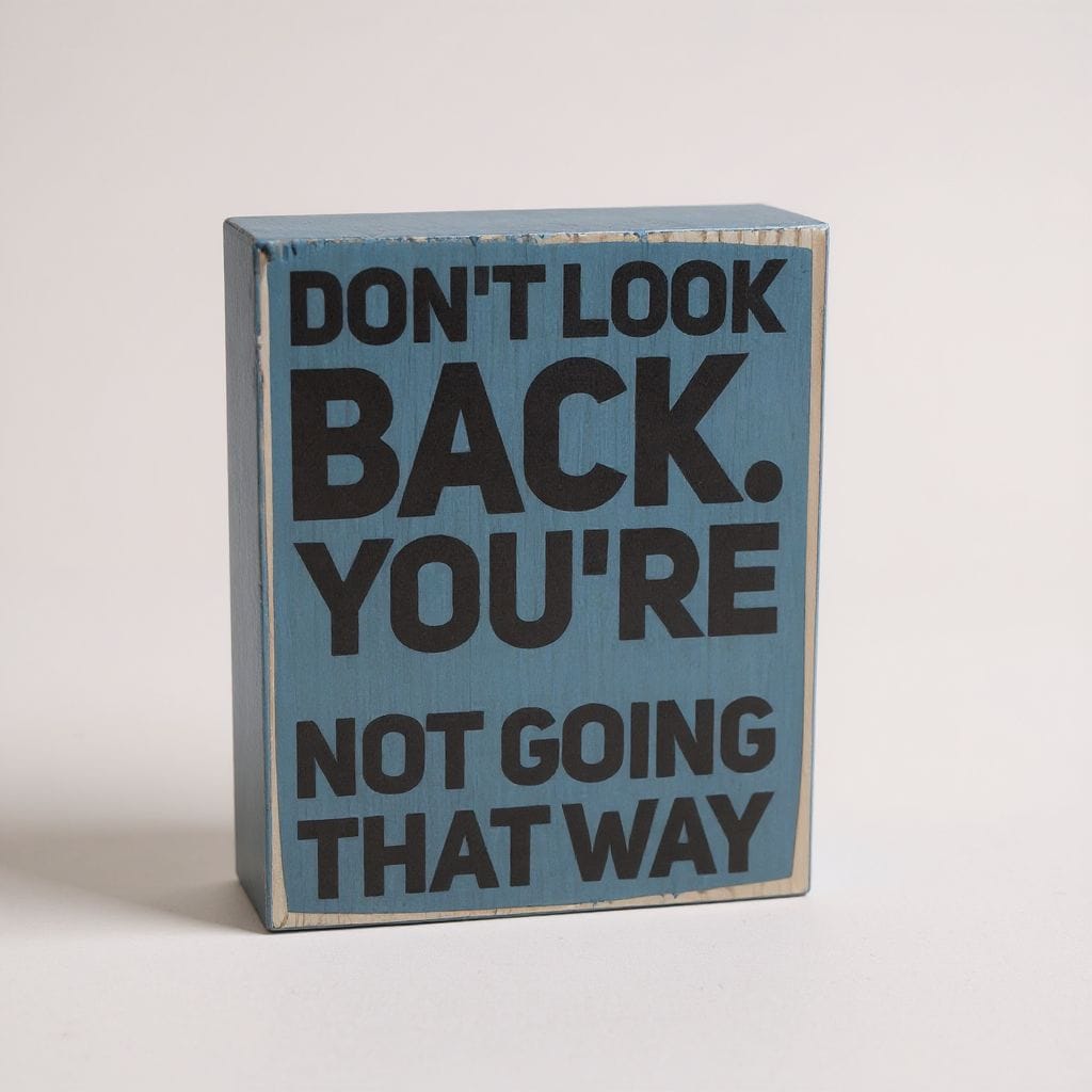 A rectangular wooden sign with a distressed light blue finish and bold black text that reads 'DON'T LOOK BACK. YOU'RE NOT GOING THAT WAY', photographed on a plain light background.