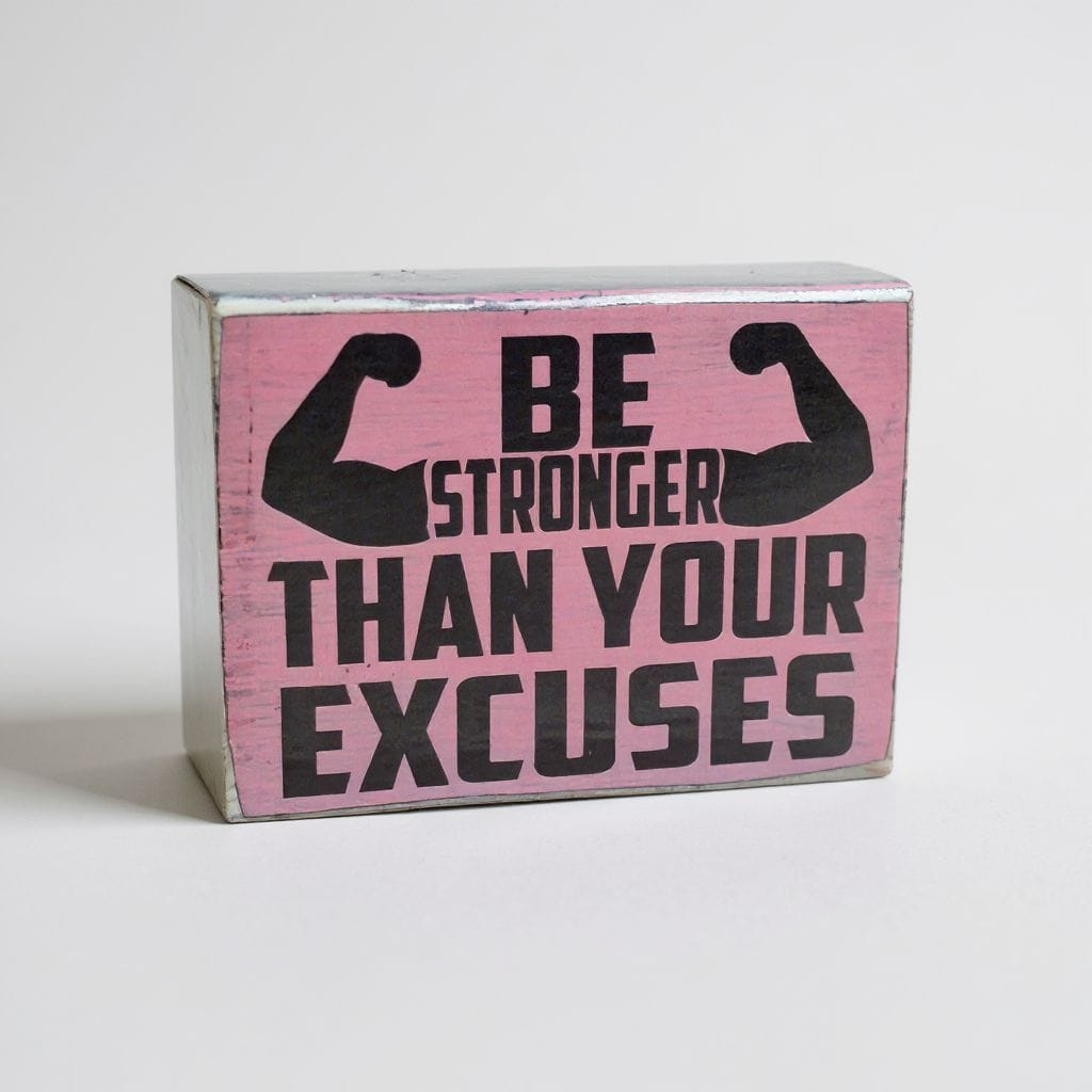 A rectangular wooden block sign with a distressed pink face and silver-gray sides, featuring the motivational quote 'BE STRONGER THAN YOUR EXCUSES' in bold black text with flexing arm graphics.