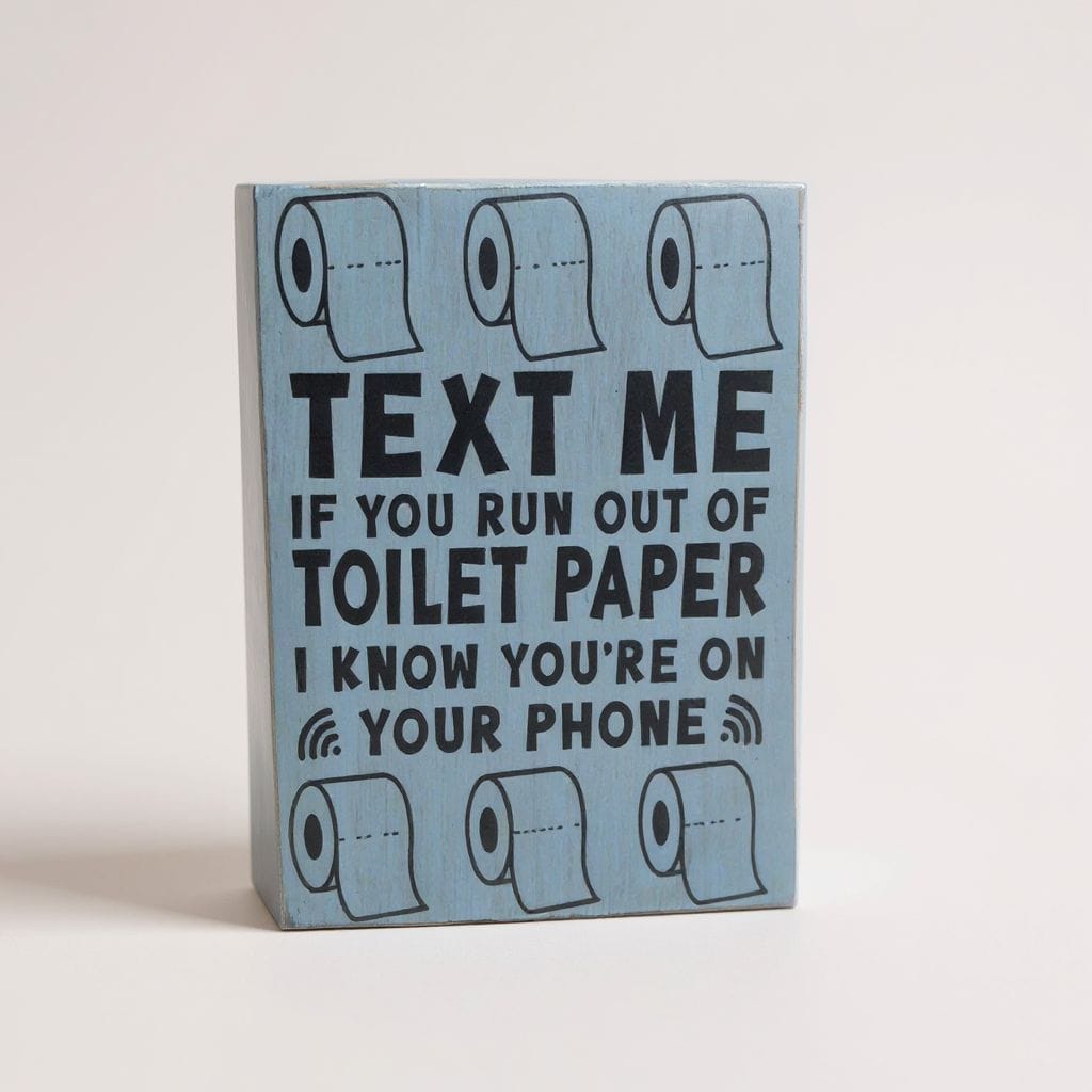 A rectangular light blue wooden sign with black text that reads 'TEXT ME IF YOU RUN OUT OF TOILET PAPER I KNOW YOU'RE ON YOUR PHONE' and toilet paper illustrations at the top and bottom, photographed against a plain light background.