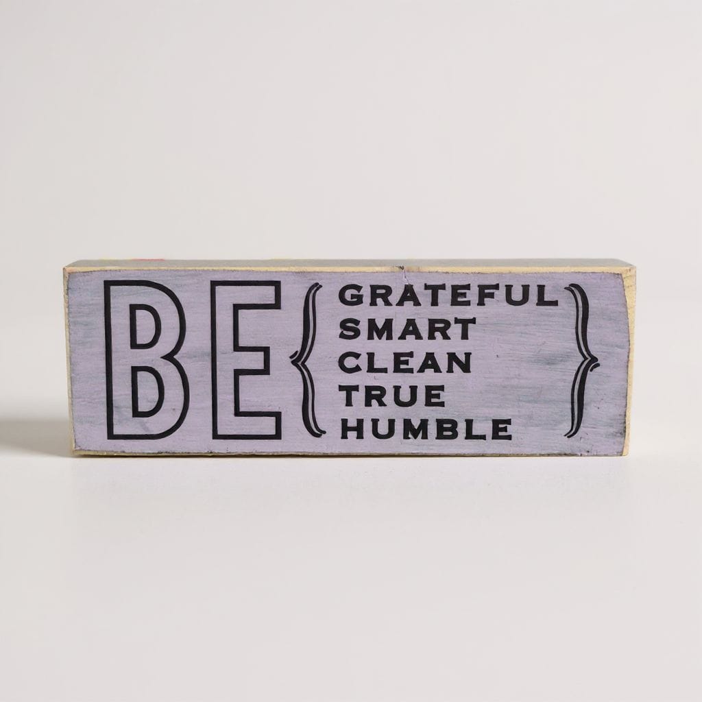 A rectangular decorative wooden sign with a distressed light lavender-gray finish. The sign features the text 'BE GRATEFUL SMART CLEAN TRUE HUMBLE' in black lettering, with 'BE' in large outlined letters and the rest enclosed in curly braces.