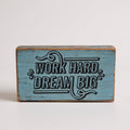 A rectangular rustic wooden block sign with a distressed light blue painted finish and black text that reads 'WORK HARD DREAM BIG' with decorative flourishes, shown on a plain light background.