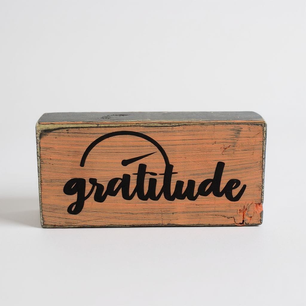 A rectangular wooden block sign with a weathered orange-brown finish, featuring the word 'gratitude' in black script under a simple black arch graphic, set against a plain light gray background.