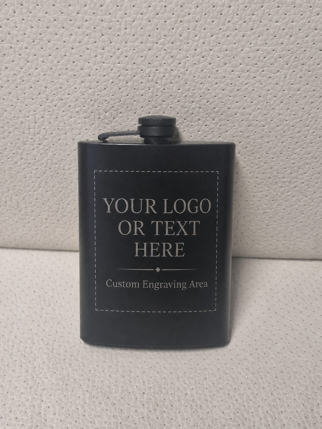 A black metal flask with a screw-top lid and a small loop handle, shown against a light textured wall. The front has a white placeholder engraving that reads 'YOUR LOGO OR TEXT HERE' and 'Custom Engraving Area'.