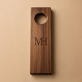 A personalized wooden door hanger or pegboard insert with a circular hole at the top and engraved initials 'MH' in the center, shown on a solid beige background.