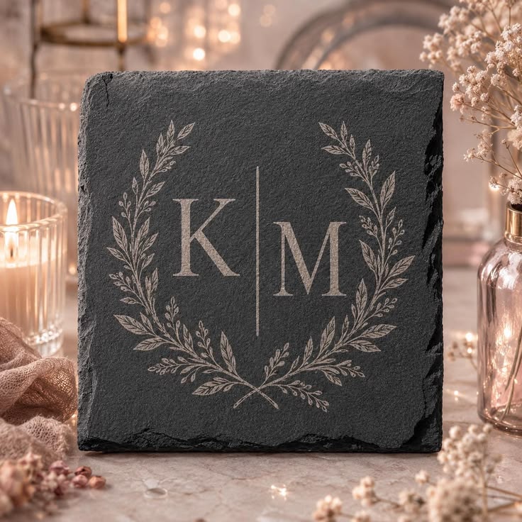 A square dark charcoal slate coaster with a personalized monogram design featuring the initials 'K' and 'M' separated by a vertical line, framed by a silver-gray laurel wreath. The coaster is styled on a tabletop with warm bokeh background lights, candles, and dried flowers.