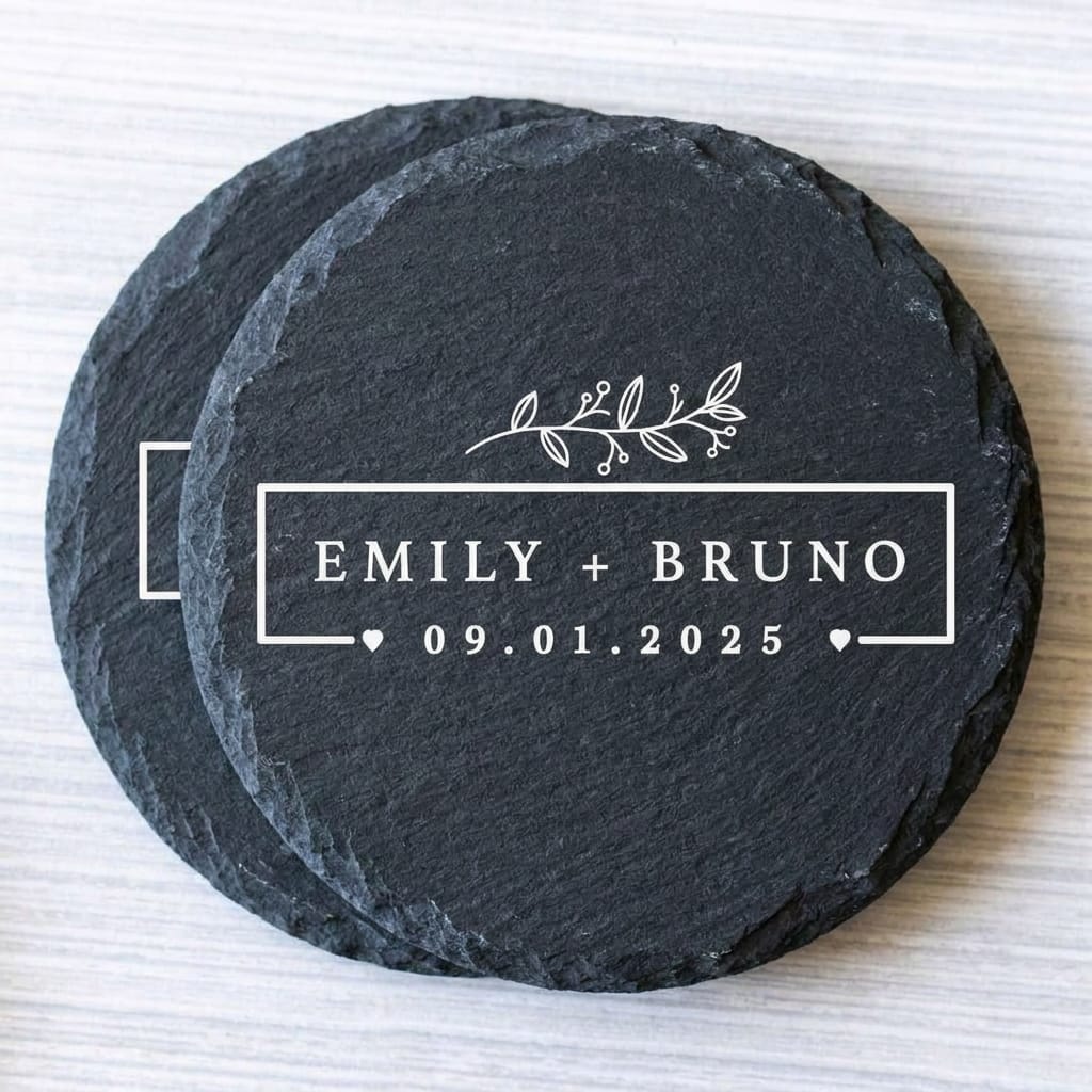 Two round black slate coasters stacked on a light wood surface, with white engraved text reading 'EMILY + BRUNO 09.01.