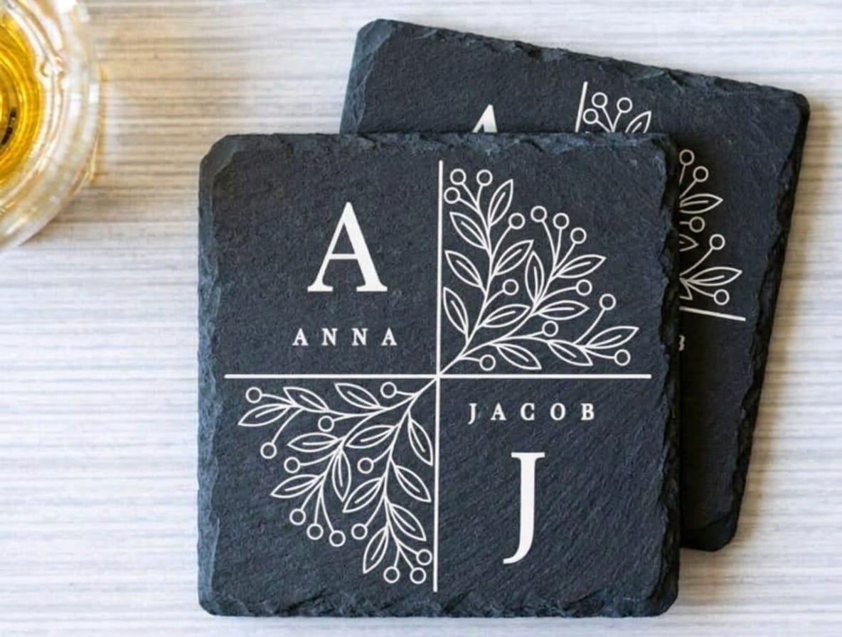 Two square black slate coasters stacked on a light wood surface. The front coaster is personalized with a white cross dividing the surface into four quadrants, featuring the monogram 'A', the name 'ANNA', botanical line art, and the monogram 'J', the name 'JACOB'.