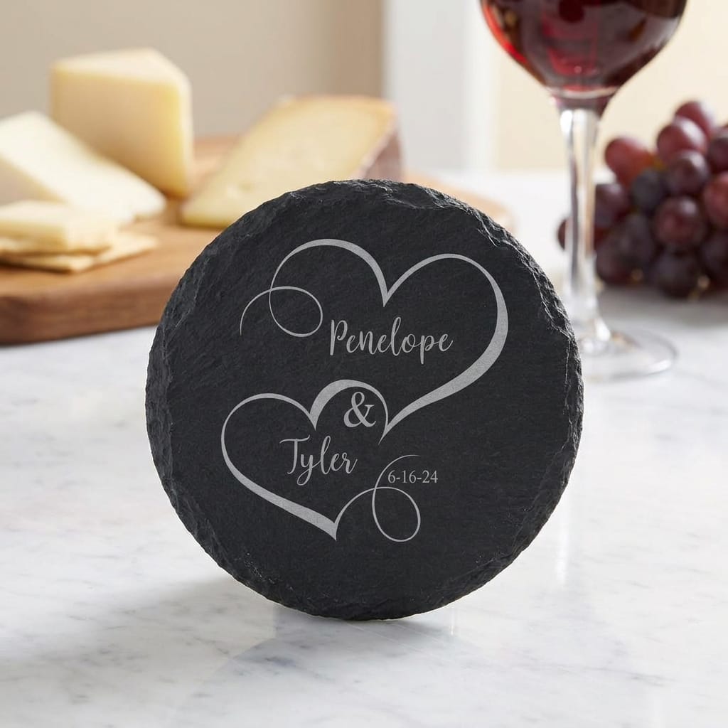 A round black slate cheese board cover engraved with two white hearts, the names 'Penclope & Tyler', and the date '6-16-24', shown on a marble surface with cheese, grapes, and a glass of red wine in the background.