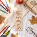 A wooden bookmark with engraved text reading 'BEST wishes to a teacher who makes learning so much easier' is shown in a styled flat-lay with colored pencils, an apple, old books, autumn leaves, and eyeglasses on a white fabric background.