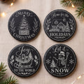 Four round black holiday coasters with white printed Christmas illustrations and phrases, arranged on a beige fabric surface with pine branches and warm string lights in the background.