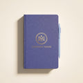 Custom navy blue hardcover journal with gold foil circular monogram logo and company name, flat lay on a clean cream background with a blue pen