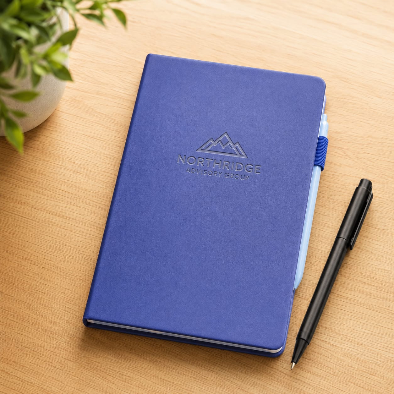 Custom navy blue vegan leather journal with debossed Northridge Advisory Group mountain logo, laying on a wooden desk next to a black pen and green plant — personalized corporate gift notebook