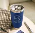 A blue insulated travel tumbler with a stainless steel rim, filled with ice, placed on a tabletop next to a patterned fabric and a pen. The tumbler has a circular mountain-and-droplet logo and the word ALPINE printed on the front.