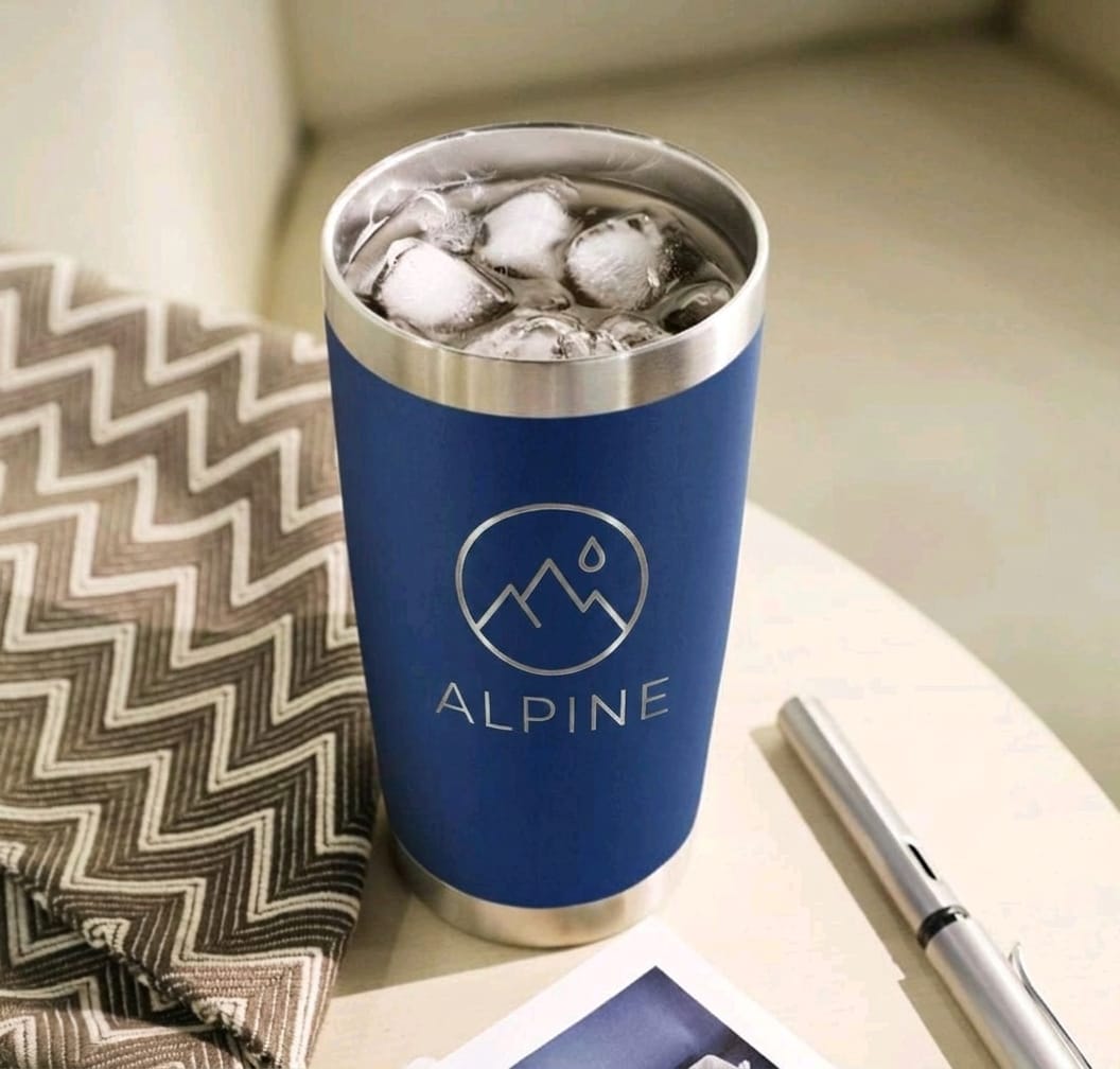 A blue insulated travel tumbler with a stainless steel rim, filled with ice, placed on a tabletop next to a patterned fabric and a pen. The tumbler has a circular mountain-and-droplet logo and the word ALPINE printed on the front.
