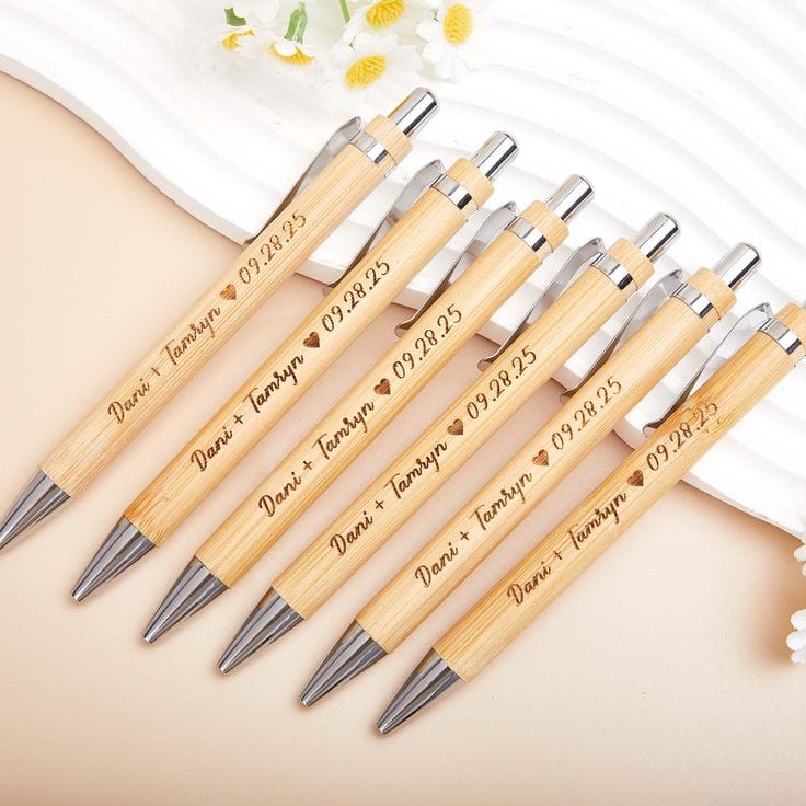 Six personalized bamboo ballpoint pens with silver metal tips and clips, engraved with names and a date, arranged diagonally on a beige surface with a white decorative plate in the background.