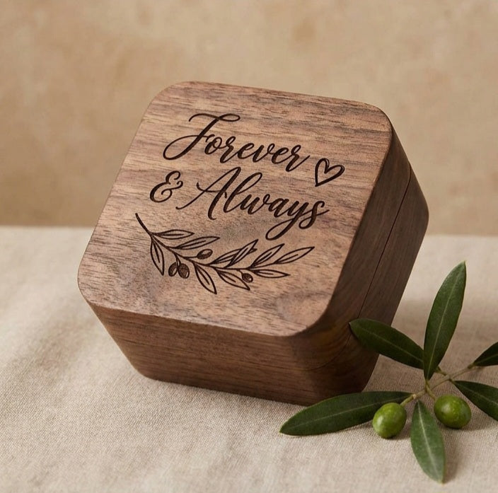 A square wooden keepsake box with rounded corners, engraved with 'Forever & Always' and an olive branch design, styled on a beige fabric surface with green olives in the foreground.