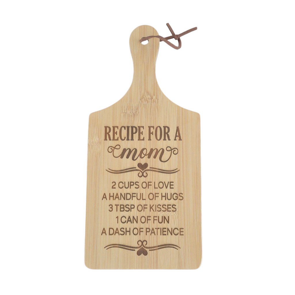 A light natural wood cutting board with a handle, featuring an engraved recipe for 'mom' with hearts and decorative flourishes. A small brown cord loop is attached at the top, and the board is photographed against a solid white background.