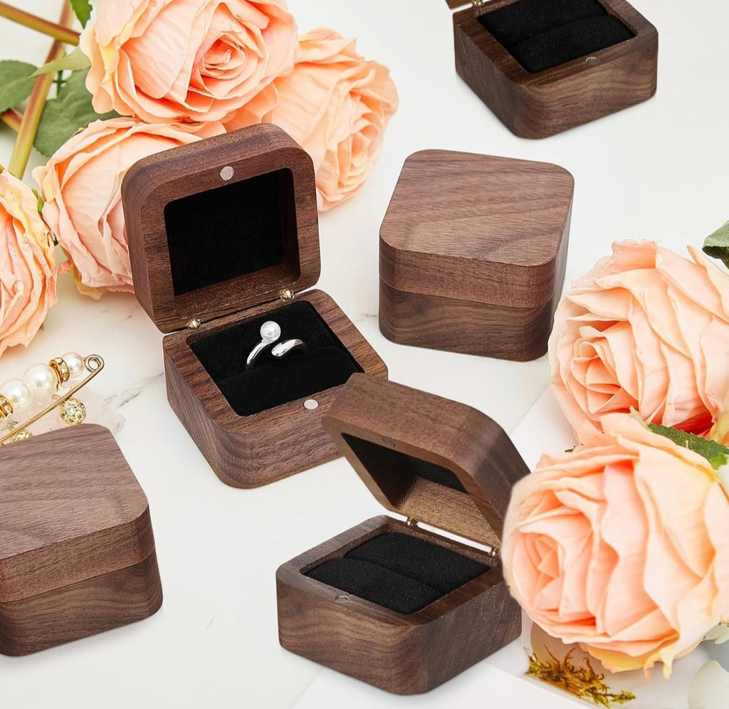 Dark brown wooden ring boxes with black velvet interiors, shown open and closed on a white surface with peach roses and a pearl pin as props.