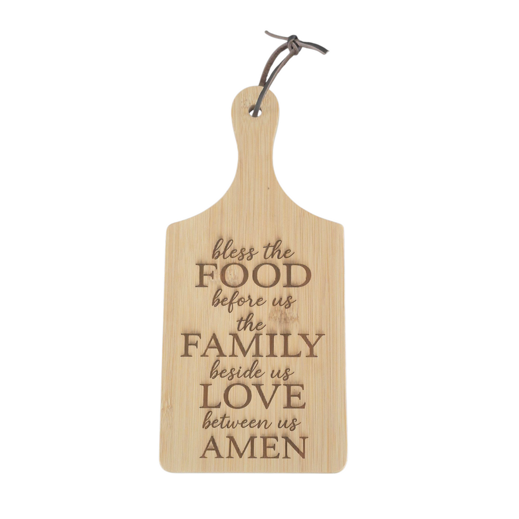 A light natural wood serving board with a handle, featuring engraved text that reads 'bless the FOOD before us the FAMILY beside us LOVE between us AMEN' in a dark brown tone. It has a metal hanging loop at the top and is shown against a plain white background.