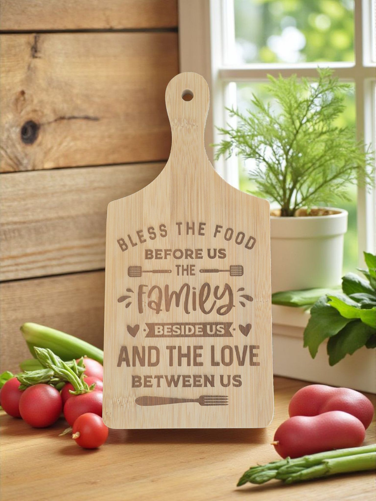 A personalized wooden cutting board with a warm natural wood finish, featuring the engraved text 'Bless the food before us the family and the love between us'. The cutting board is styled on a wooden table with fresh vegetables (radishes, asparagus) and a potted plant in the background, creating a cozy kitchen atmosphere.