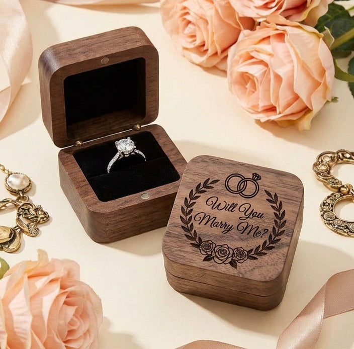 A wooden engagement ring box with one open box showing a silver ring on a black velvet insert and one closed box engraved with 'Will You marry me?' and a ring motif, styled with peach roses and gold jewelry on a cream surface.